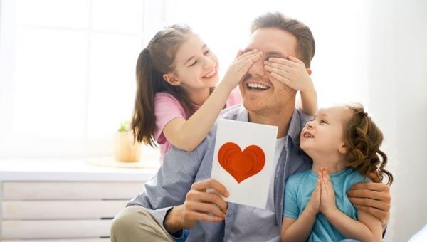 Ilustračné foto: Muž má v ruke výkres so srdcom, dcéry mu zakrývajú oči. Všetci traja sa smejú. Zdroj: iStock