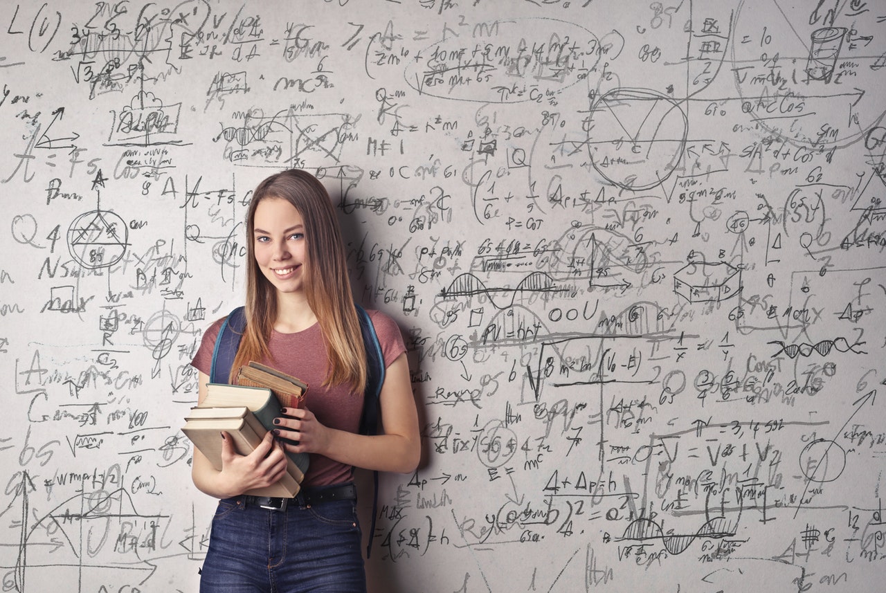 Ilustračný obrázok: Študentka s knihami pred tabuľou popísanou rovnicami. Zdroj: Pexels.com